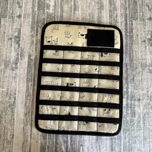 May include: A rectangular, quilted organizer with a cream-colored background featuring a repeating pattern of black and white dog illustrations. The organizer has black horizontal straps and a black border, with a black velcro patch.