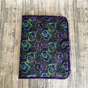 May include: A rectangular fabric notebook cover featuring a dark purple and black background with a repeating pattern of swirling, colorful abstract shapes in shades of neon green, blue, and yellow. The cover includes a small fabric tab closure.