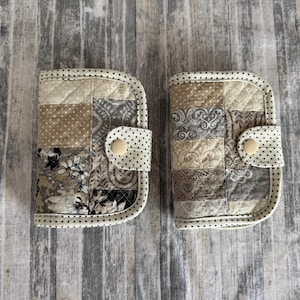 May include: Two quilted fabric wallets with a patchwork design in neutral tones of beige, grey, and brown. Each wallet has a rounded flap with a polka dot pattern and a button closure. The edges are trimmed with black polka dots.