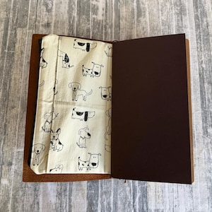 May include: Open brown leather journal with a cream-colored fabric lining featuring a repeating black and white dog illustration. The right page is a solid dark brown color.