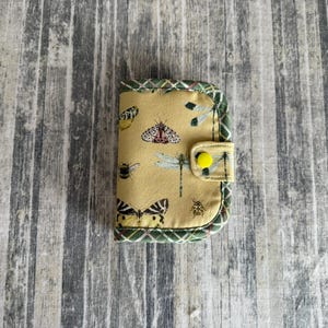 May include: A small, fabric wallet with a yellow background and insect illustrations. The wallet features a green and white checkered border and a yellow button closure. Butterflies, bees, and dragonflies are depicted on the wallet.