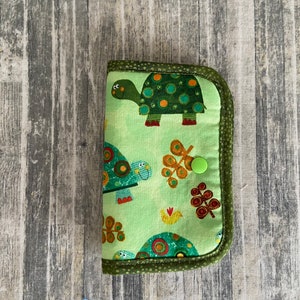 May include: A green fabric zippered pouch with a turtle design. The pouch has a green button closure and a green and brown trim.