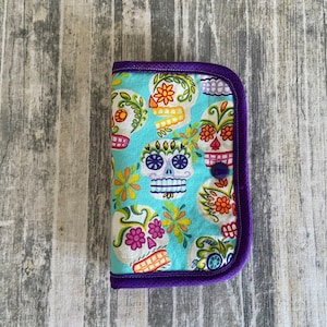 May include: A fabric case with a turquoise background featuring colorful sugar skull designs. The case is trimmed with purple fabric and a matching button closure. The sugar skulls have floral and decorative elements.