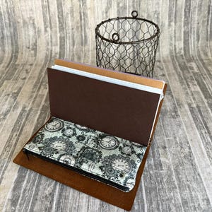 May include: Open brown leather journal with a decorative clock face pattern on the inside cover. The journal is open, revealing several pages of different colors. A wire basket sits in the background.