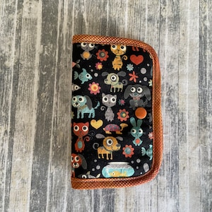 May include: A black fabric wallet with a brown trim and a colourful pattern of cartoon animals, including cats, dogs, and fish. The wallet has a button closure.