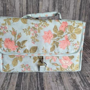 May include: A floral print handbag with a light blue background and pink roses. The bag has a handle and a silver-colored clasp. The bag is rectangular with rounded edges and a flap closure.