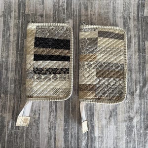 May include: Two quilted zippered pouches in neutral tones. One pouch features a patchwork design with black, beige, and cream fabrics. The other has a similar patchwork design in shades of beige and brown. Both have white zipper pulls.