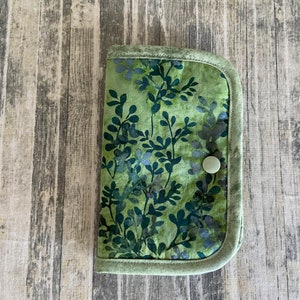 May include: A green fabric pouch with a floral pattern and a white button closure. The pouch is on a woodgrain background.