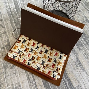 May include: Open brown leather journal with a cream-coloured cat print zippered pouch. The journal has a dark brown interior and a notebook with white pages. A black wire basket is in the background.
