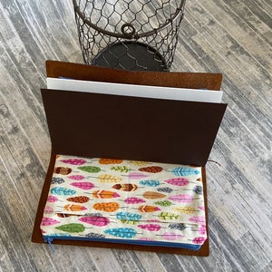 May include: A brown leather notebook with a zippered pocket. The pocket is lined with a fabric featuring a colorful feather pattern.