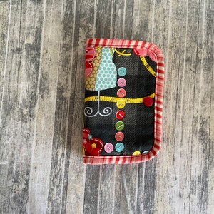 May include: A small black fabric case with a red and white trim. The case has a sewing theme with buttons, a measuring tape, and a sewing machine on the front.