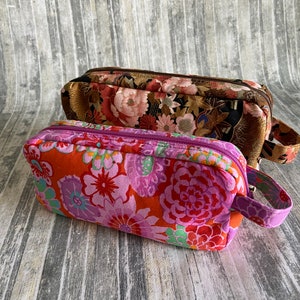 May include: Two floral patterned zippered pouches with handles. The top pouch is brown with gold accents and pink and white flowers. The bottom pouch is red with pink, white, and green flowers.