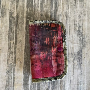 May include: A small, rectangular case with a curved top, featuring a dark red and black abstract design with text. The case is trimmed with a patterned border and secured with a purple button.