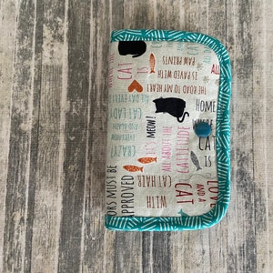 May include: A teal and white fabric wallet with a cat theme. The wallet has a snap closure and features a variety of cat-related phrases, including "Cat is Love and a Cattitude", "All About the Cat Lady", and "The Road to My Heart is Paved with Paw Prints".
