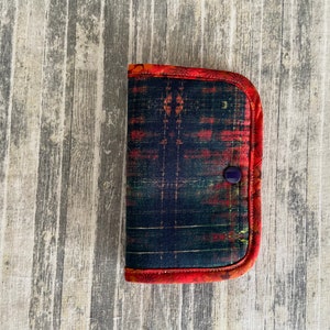 May include: A small, red and black fabric wallet with a button closure. The wallet has a geometric pattern and is made of a soft, quilted fabric.