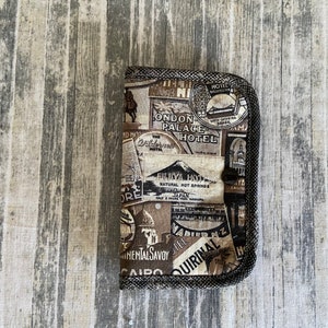 May include: A brown fabric wallet with a zipper closure. The wallet is decorated with vintage travel posters featuring images of hotels, landmarks, and cities. The text on the posters includes "London Palace Hotel", "Fujiya Hotel", "Natural Hot Springs", "Japan", "Cairo", "The Quirinal", "Wadiernz", and "Continental Savoy".