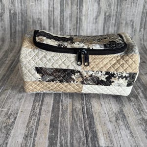May include: A quilted cosmetic bag with a black zipper closure. The bag features a patchwork design with various neutral-toned fabrics, including beige, cream, and black floral patterns. The bag is rectangular with a curved top.
