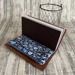 May include: A brown leather journal with a blue and black floral zippered pouch. The journal is open to reveal lined pages.