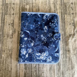May include: A blue floral fabric journal cover with a light blue border and button closure. The cover features a pattern of blue and white flowers and foliage. The journal cover is rectangular and appears to be made of fabric.