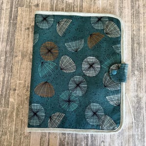 May include: A rectangular fabric folder with a teal background featuring a repeating pattern of dandelion-like seed heads in black, white, and brown. The folder has light gray piping along the edges and a small tab closure with a blue snap button.