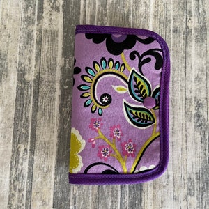 May include: A purple fabric wallet with a floral pattern in shades of green, yellow, black, and pink. The wallet has a purple button closure.