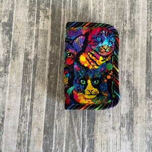 May include: A black fabric wallet with a colourful print of cats. The print features a variety of colours, including red, yellow, blue, green, and black. The wallet has a black zipper and a green button.
