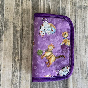May include: Purple fabric wallet with a cat design. The wallet has a purple button closure and a purple trim.