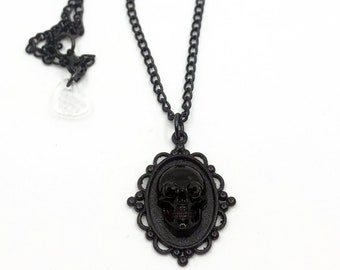 May include: A black skull pendant necklace. The pendant features a black skull within an oval frame with decorative edges. The necklace chain is also black. The necklace is displayed against a white background.