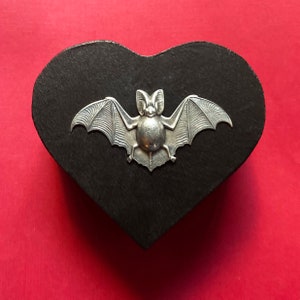 Könnte beinhalten: Eine schwarze herzförmige Holzkiste mit einem silbernen Fledermaus-Design auf der Vorderseite.