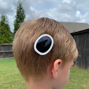 Puede incluir: Una pegatina ovalada blanca con un círculo negro en el centro. La pegatina está pegada a la cabeza de una persona.