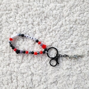 May include: A beaded keychain with the word "GHOST" spelled out in white letter beads. The keychain features red, black, and silver beads, with a star charm and a small gun charm.