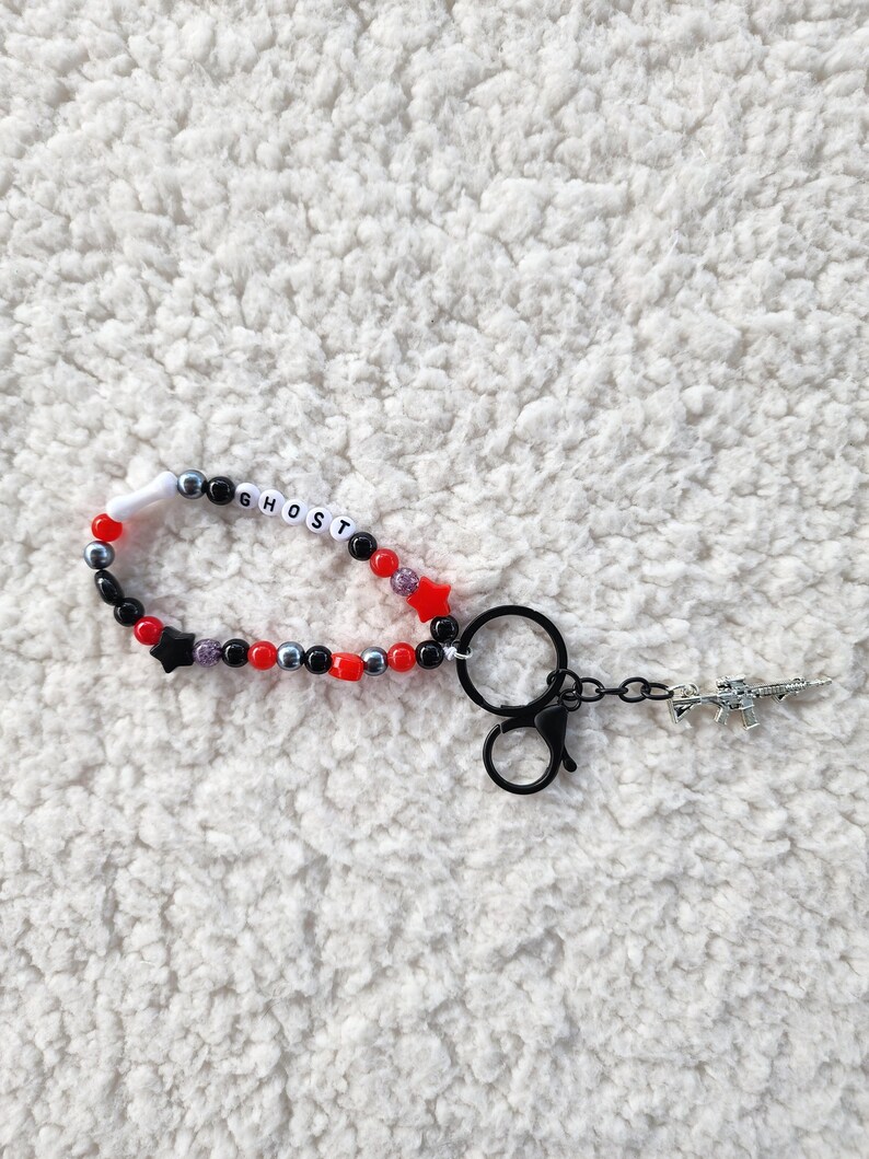 May include: A beaded keychain with the word "GHOST" spelled out in white letter beads. The keychain features red, black, and silver beads, with a star charm and a small gun charm.