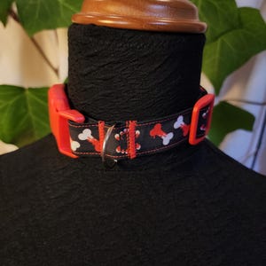 Può includere: Un collare nero per cani con un motivo a ossa rosse e bianche. Il collare ha una fibbia in plastica rossa e un anello a D in metallo argentato. Il collare è esposto su una testa di manichino nera.