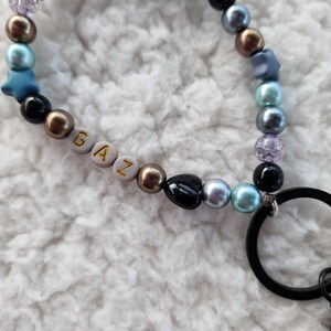 May include: A beaded keychain wristlet with a mix of pearl and star-shaped beads in black, blue, gold, and iridescent colors. The word "GAZ" is spelled out with gold letters on white beads. A black metal ring is attached.