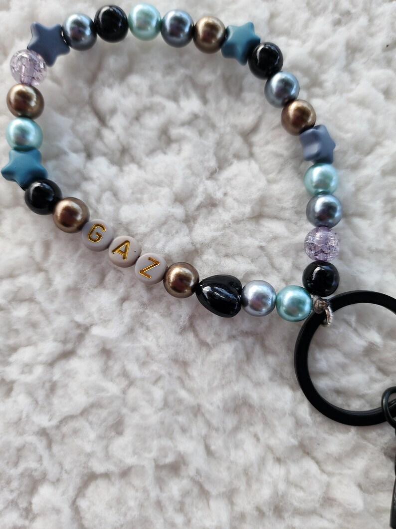 May include: A beaded keychain wristlet with a mix of pearl and star-shaped beads in black, blue, gold, and iridescent colors. The word "GAZ" is spelled out with gold letters on white beads. A black metal ring is attached.