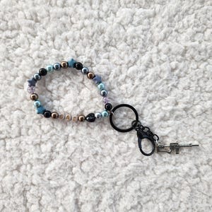 May include: A beaded keychain with a variety of beads in colors like blue, gold, and black. The keychain includes a charm that resembles a small gun. The word "GAZ" is spelled out with letter beads.