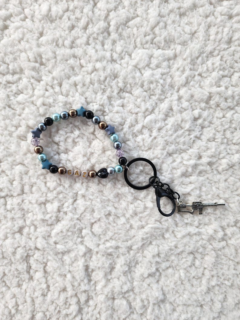 May include: A beaded keychain with a variety of beads in colors like blue, gold, and black. The keychain includes a charm that resembles a small gun. The word "GAZ" is spelled out with letter beads.
