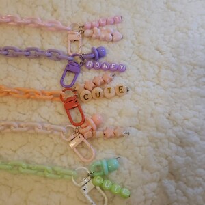Op de afbeelding: Vijf kleurrijke plastic kettingen met bedels en letterkraaltjes. De kettingen zijn roze, paars, oranje, wit en groen. De bedels omvatten een speen, sterren en harten. De letterkraaltjes spellen "SWEET", "HONEY", "CUTE" en "CUTE" uit.
