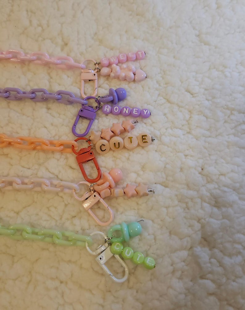Op de afbeelding: Vijf kleurrijke plastic kettingen met bedels en letterkraaltjes. De kettingen zijn roze, paars, oranje, wit en groen. De bedels omvatten een speen, sterren en harten. De letterkraaltjes spellen "SWEET", "HONEY", "CUTE" en "CUTE" uit.