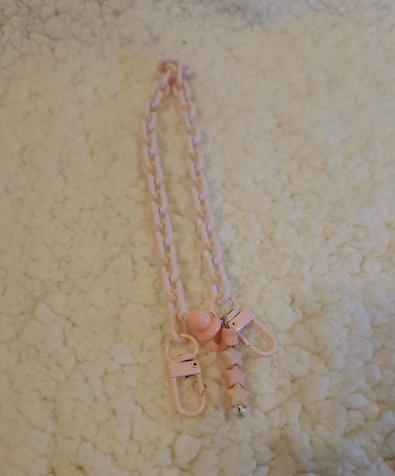 Op de afbeelding: Een roze plastic ketting met twee roze clips en een roze kralenbedel. De ketting is gemaakt van in elkaar grijpende schakels en de bedel is een reeks stervormige kralen.