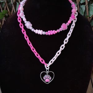 May include: A two-strand necklace with pink and white components. The top strand has pink flower and bead details. The bottom strand is a pink chain and a white chain. A silver heart pendant with a pink paw print hangs from the bottom chain.
