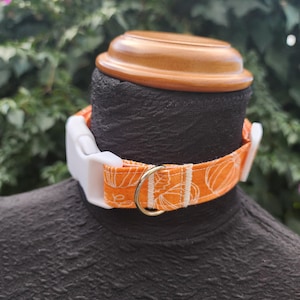 May include: An orange and white pumpkin-themed dog collar with a white plastic buckle and a gold-colored metal ring. The collar is on a black mannequin head, with a wooden top in the background.