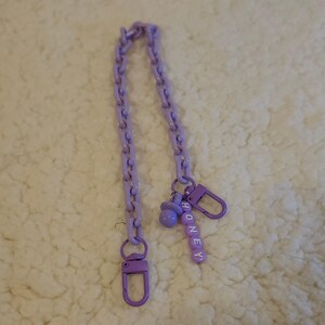 Op de afbeelding: Een paarse ketting met twee sluitingen en een bedel met de tekst "HONEY" in witte letters.