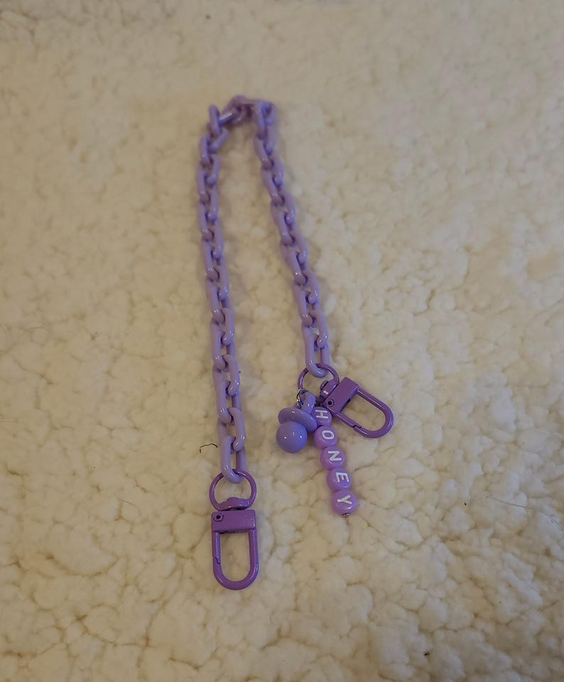 Op de afbeelding: Een paarse ketting met twee sluitingen en een bedel met de tekst "HONEY" in witte letters.