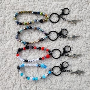 May include: Four beaded keychains with black clasps and small gun charms. The keychains feature various colors of beads, including gold, silver, black, red, white, and blue. Some keychains have letter beads spelling out words.