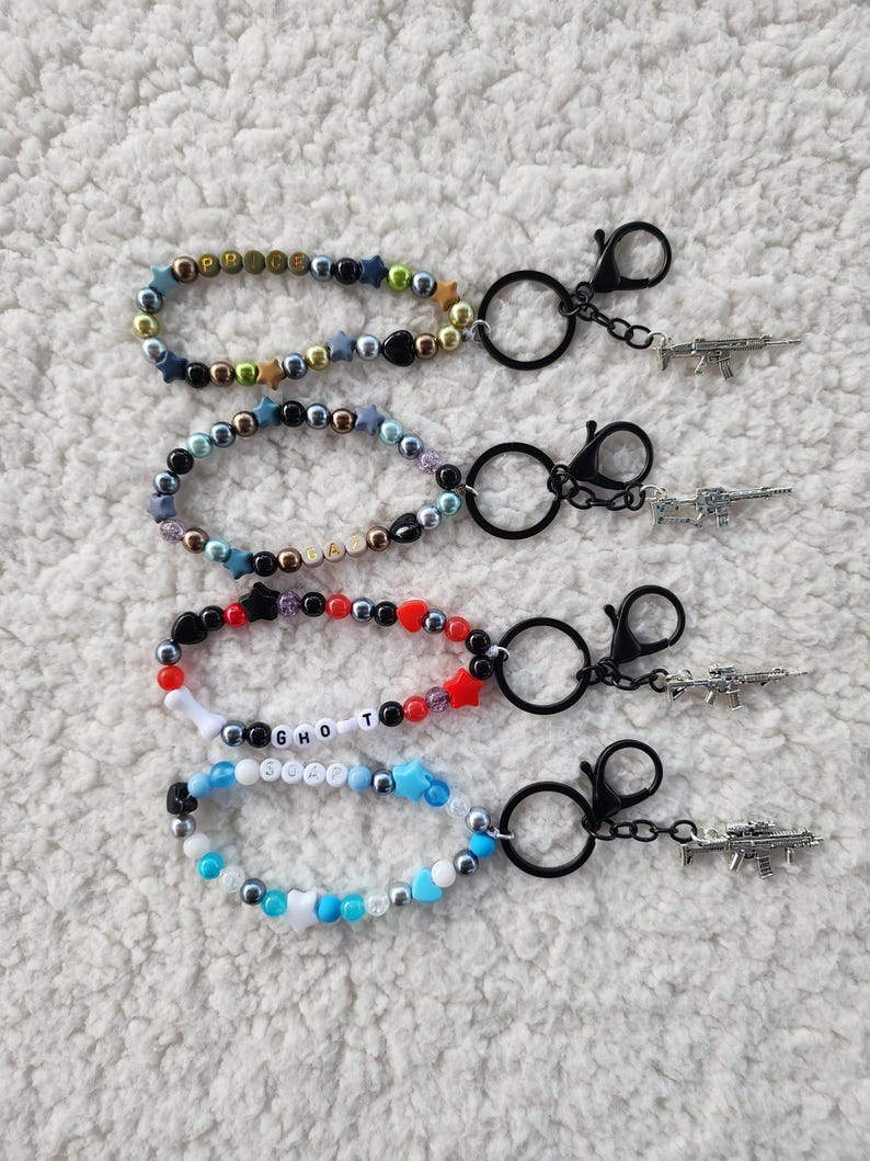 May include: Four beaded keychains with black clasps and small gun charms. The keychains feature various colors of beads, including gold, silver, black, red, white, and blue. Some keychains have letter beads spelling out words.
