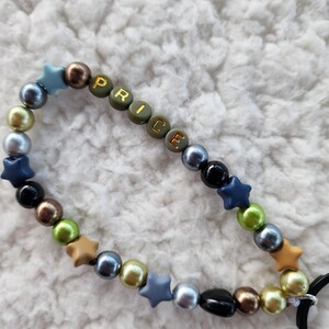 May include: A beaded accessory with the word "PRICE" spelled out in olive green letter beads. The accessory features a mix of colorful beads, including blue, gold, black, and brown, and star-shaped beads. A black loop is attached.