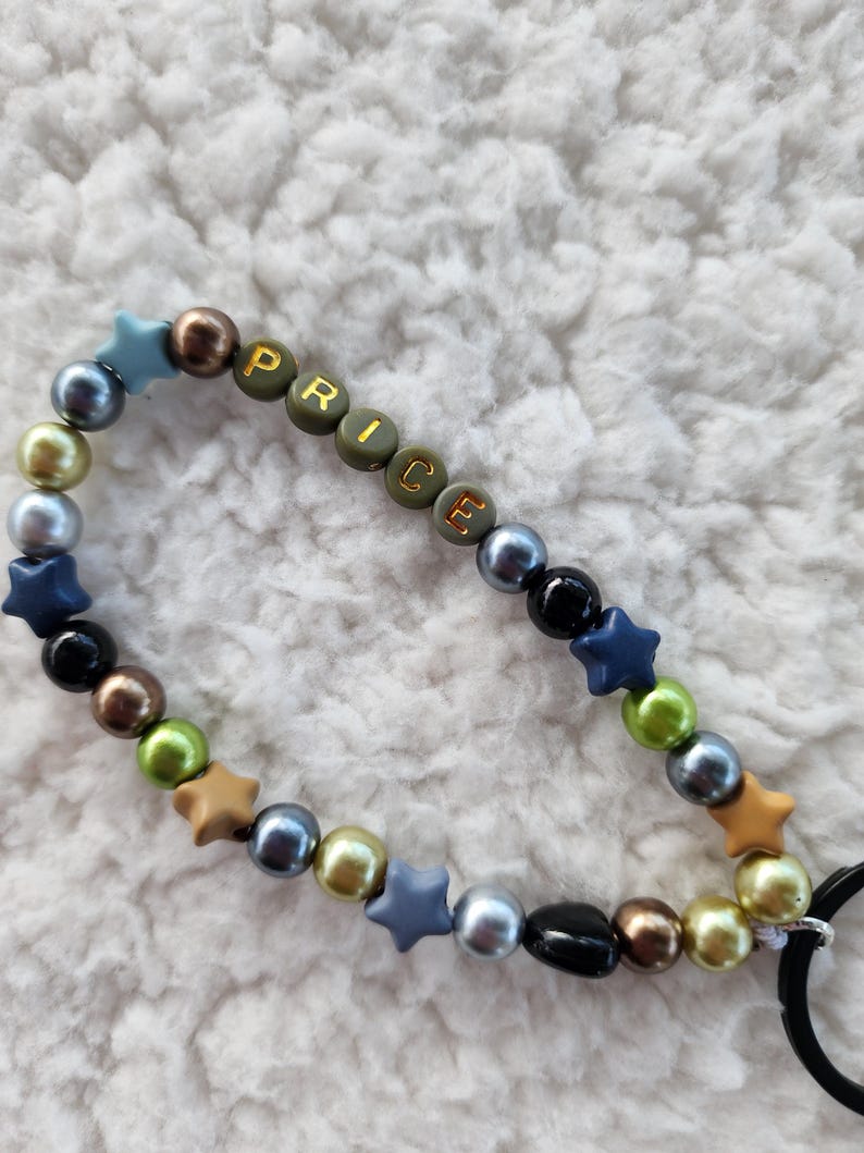 May include: A beaded accessory with the word "PRICE" spelled out in olive green letter beads. The accessory features a mix of colorful beads, including blue, gold, black, and brown, and star-shaped beads. A black loop is attached.