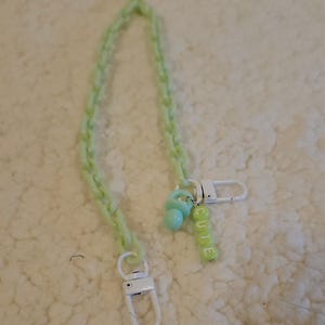 Op de afbeelding: Een lichtgroene plastic ketting met een witte sluiting en een lichtgroene speenbedel. De bedel heeft een zilveren ring met het woord "CUTE" gespeld in lichtgroene kralen.