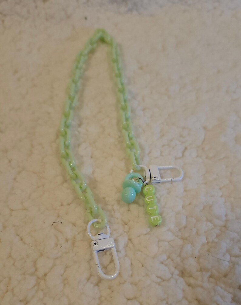 Op de afbeelding: Een lichtgroene plastic ketting met een witte sluiting en een lichtgroene speenbedel. De bedel heeft een zilveren ring met het woord "CUTE" gespeld in lichtgroene kralen.
